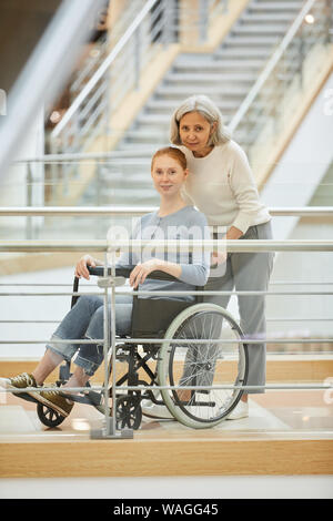 Ritratto di infermiere senior in piedi vicino alla giovane donna disabile in sedia a rotelle e che guardando la fotocamera all'ospedale Foto Stock