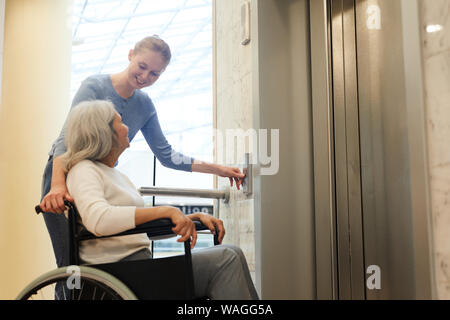 Giovane donna premendo il pulsante di ascensore e sorridente al senior donna disabile mentre era seduto nella sedia a rotelle Foto Stock