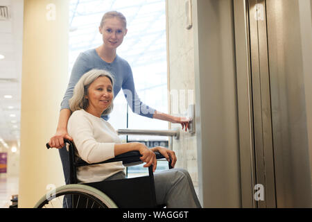 Ritratto di giovane infermiere senior con paziente in carrozzella sorridente in telecamera durante l'attesa per l'ascensore all'ospedale Foto Stock