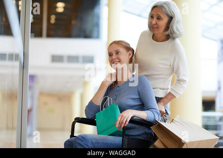 Senior donna insieme con la giovane donna sorridente in sedia a rotelle di trascorrere del tempo insieme nel centro commerciale per lo shopping Foto Stock