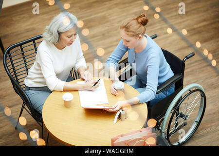 Donna Senior il riempimento di un qualche documento con la giovane donna in sedia a rotelle con tavoletta digitale mentre sono seduti a tavola in cafe Foto Stock