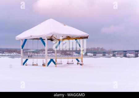 Gazebo abbandonato per l'inverno con banchi di oscillazione tra i cumuli di neve sulla riva del fiume Foto Stock