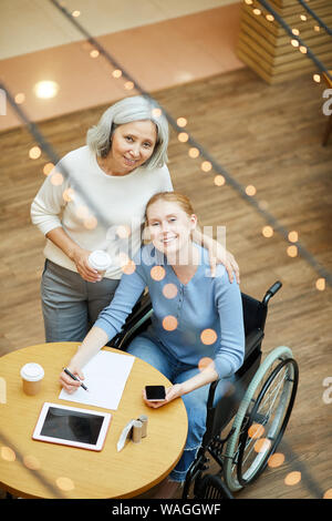 Ritratto di donna senior e la giovane donna disabile in carrozzella sorridente in telecamera durante il riempimento il documento al tavolo in cafe Foto Stock