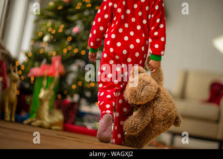 Ragazza giovane che porta il suo orsacchiotto verso l'albero di Natale la mattina di Natale. Foto Stock