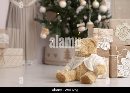 Chiudere su toy Teddy bear nei pressi della montagna di doni confezionate in carta artigianale su uno sfondo sfocato di un albero di Natale decorato, il fuoco selettivo Foto Stock
