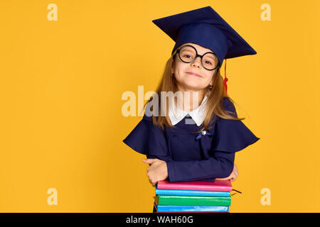 Allegro schoolgirl in abito di graduazione sorridere mentre appoggiato sulla pila di libri colorati su sfondo giallo Foto Stock