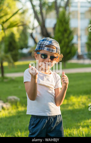 Ritratto di un bambino in posa di un parco vestito in abiti estivi, cappello e occhiali da sole, rendendo il pollice in alto gesto. Foto Stock