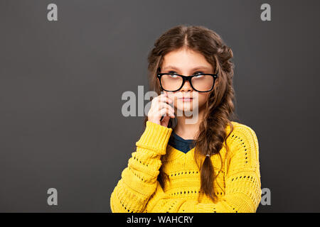 Pensiero serio smorfie schoolgirl in occhiali cercando su grigio di sfondo per studio. Si torna a scuola. Il concetto di istruzione. Primo piano Foto Stock