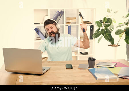 Odio di ufficio di routine. Uomo Barbuto guy ufficio cuffie swing martello sul computer. Connessione a internet lenta. Software obsoleto. Computer lag. Ragioni per computer in ritardo. Come fissare il lento sistema di ritardo. Foto Stock