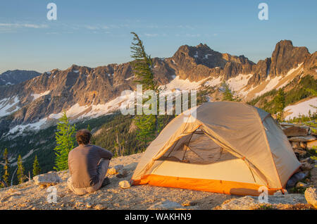 Uomo adulto allo spuntar del giorno seduto accanto a backpacking tenda e guardando a vista del picco spietato sulla cresta sopra Tagliagole Pass, vicino Pacific Crest Trail. North Cascades, nello Stato di Washington Foto Stock