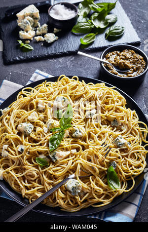 Delizioso Spaghetti con pesto di basilico e formaggio blu su una piastra nera su una tavola di cemento con il pesto in una ciotola nero e gli ingredienti su un taglio di ardesia Foto Stock