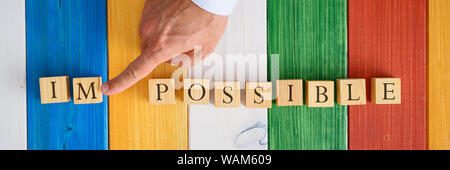 Ampia vista immagine del dito maschio spingendo lontano sigla IM dalla parola impossibile scritto con cubetti di legno su sfondo colorato. Foto Stock