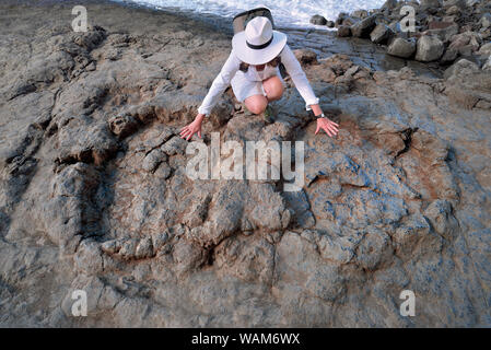Donna con cappello bianco dirigere le mani per due enormi tracce di dinosauri in una roccia sul mare Foto Stock