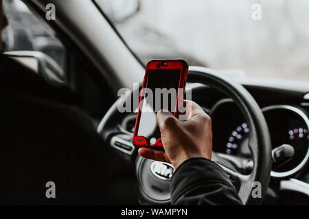 Una persona che tiene uno smartphone rosso seduto davanti al volante di un veicolo Foto Stock