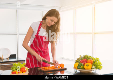 Giovani caucasici donna, capelli biondi, capelli lunghi, indossa una t-shirt bianco e rosso permanente della catenaria, affettare le carote sulla tabella alla moderna cucina bianca su Foto Stock