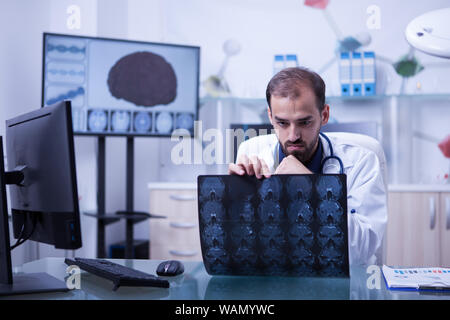 Bello barbuto medico guardando una radiografia del cervello. Medico seduto alla sua scrivania in un armadio di controllo di un cervello scansione tc. Foto Stock