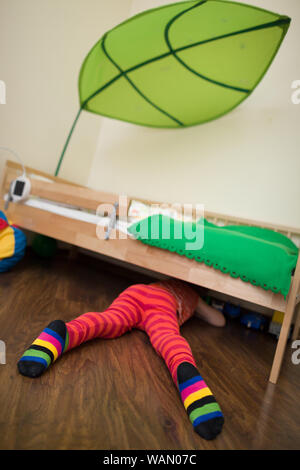 Little Boy in rosso pantalone a righe e le calze guardando sotto il letto. Toy sotto il letto. Letto matrimoniale con baldacchino nella stanza dei bambini. Foto Stock