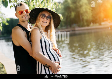 Foto di donna incinta in hat e gli uomini in rosa bicchieri in piedi sulla riva del fiume sul giorno di estate Foto Stock