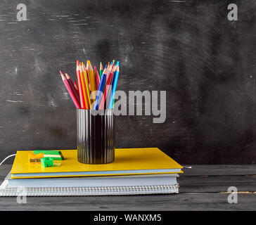 Pila di notebook, cancelleria nero con vetro multi-colore matite in legno sullo sfondo di un vuoto di gesso nero bordo, torna al concetto di scuola Foto Stock