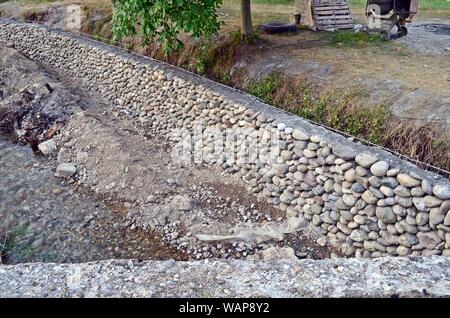 Un muro di pietra a riva del fiume in un villaggio di montagna nel nord del Montenegro; con un mescolatore di cemento, pallet di legno e un cucchiaio di metallo in background Foto Stock