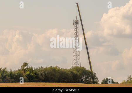 Un 5G torre di trasmissione è eretta su un campo con l aiuto di un enorme gru Foto Stock