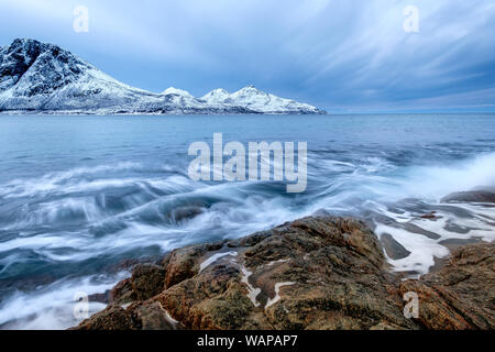 La frantumazione delle onde sulle rocce in inverno con cime innevate sullo sfondo, KvaløyaTromsø Norvegia Foto Stock