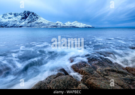 La frantumazione delle onde sulle rocce in inverno con cime innevate sullo sfondo, KvaløyaTromsø Norvegia Foto Stock