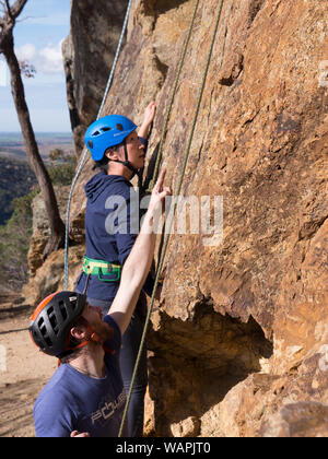 Tecnico istruttore di arrampicata, belaying un principiante alpinista e mostra paura, nervoso scalatore dove mettere le mani e i piedi sulla roccia in Australia. Foto Stock