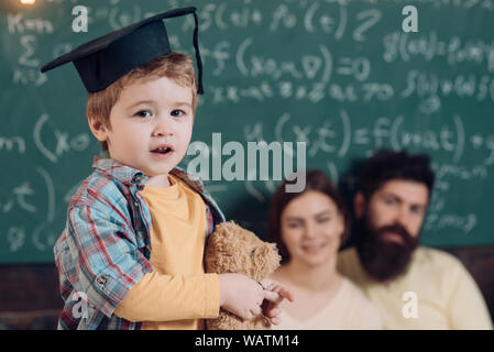 Concetto di supporto. Kid detiene orsacchiotto e di eseguire la. I genitori in ascolto il loro figlio, lavagna su sfondo. Smart bambino nel cappuccio laureati come per Foto Stock