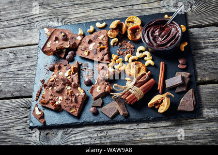 Barra di cioccolato con fichi secchi e anacardi riempimento suddivisi su pezzi in una pietra nera vassoio con ingredienti, vista orizzontale dal di sopra, close-up Foto Stock