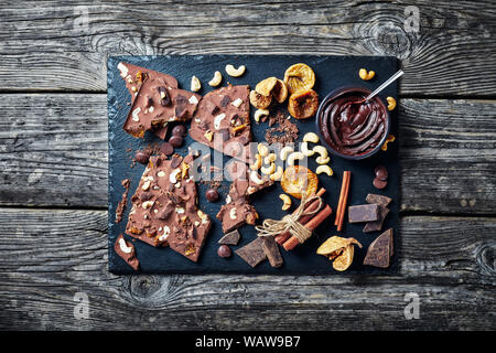 Cioccolato artigianale bar con fichi secchi e anacardi riempimento su una pietra nera vassoio con ingredienti, vista orizzontale dal di sopra, flatlay Foto Stock