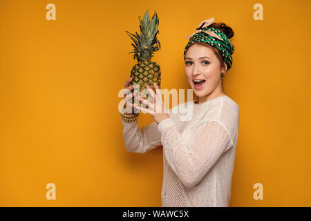 Ragazza in un maglione leggero e annodati con una sciarpa di colore la sua testa è sorridente su sfondo giallo e tenendo un ananas. Foto Stock