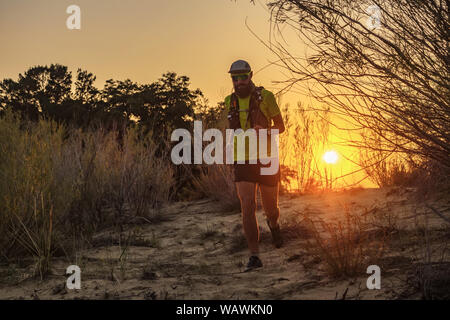 Atleta corre all'aperto in serata. Uomo in una T-shirt e shorts sullo sfondo di un tramonto nel selvaggio terreno sabbioso. Foto Stock