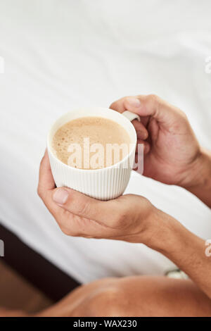 Primo piano di un giovane uomo caucasico avente un caffè in una camera da letto, accanto al letto disfatto, con biancheria bianca Foto Stock