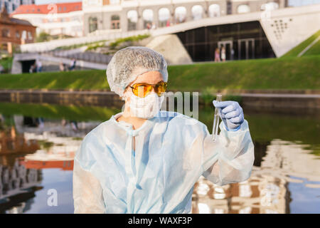 Ecologista femmina in una maschera protettiva guardando la telecamera tenendo un tubo di prova con acqua da un fiume della città Foto Stock