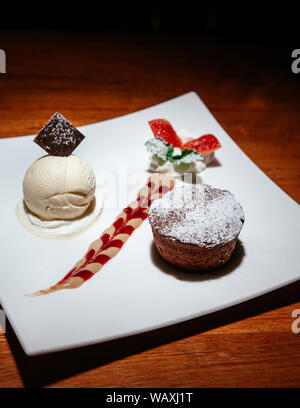 Francese dessert. Il cioccolato fondente torta di lava con gelato alla vaniglia, salsa cremosa e fragole fresche sulla panna montata. Bianco piatto da dessert su legno Foto Stock