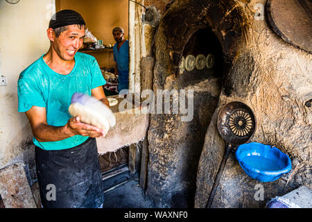 Un panettiere modella un pezzo di impasto per il pane piatto, Non (Naan) tagico, davanti a un forno di argilla tradizionale, un secondo panettiere prepara altra pasta sullo sfondo, Osh, Kirghizistan. Foto Stock