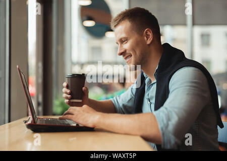 Bel giovane uomo sorridente che lavora in modalità remota nel suo computer portatile holdscup di caffè caldo in cafe Foto Stock