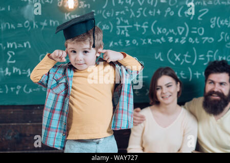 Wunderkind concetto. Smart bambino, wunderkind nel cappuccio di laurea rivolto alla sua testa. I genitori in ascolto il loro figlio, lavagna su sfondo. Ragazzo Foto Stock