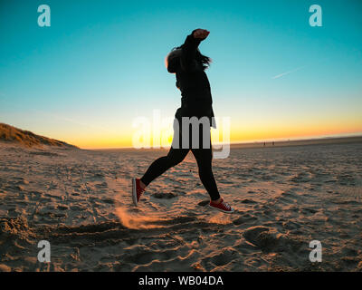 Donna che indossa cappotto e pantaloni neri saltando al tramonto sulla spiaggia Foto Stock