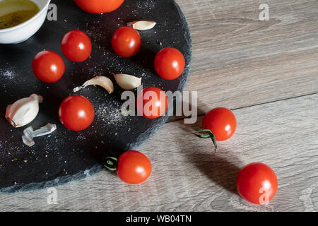 Alcuni pomodori, spicchi di aglio e un vaso con olio di oliva su una pietra nera piastra e un tavolo in legno Foto Stock
