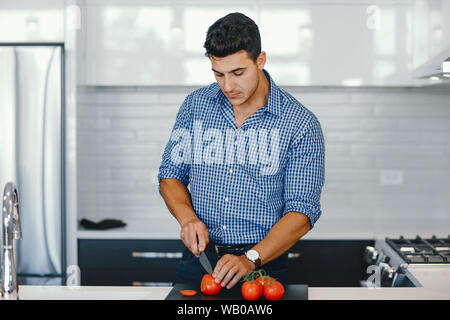 Uomo in una cucina. Un uomo bello tagliare i pomodori con un coltello. Maschio a casa con vagatables. Foto Stock