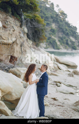 Giovane e bella con i capelli lunghi sposa in abito bianco con il suo giovane marito sulla spiaggia vicino a grosse pietre Foto Stock