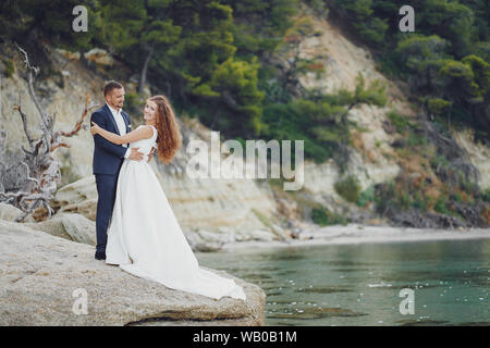 Giovane e bella con i capelli lunghi sposa in abito bianco con il suo giovane marito vicino al fiume Foto Stock