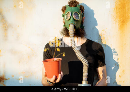 Ritratto di un triste uomo con maschera a gas azienda vaso essiccato sul grigio Sfondo arrugginito. Concetto di poveri ecologia, inquinamento atmosferico e rischi delle radiazioni Foto Stock