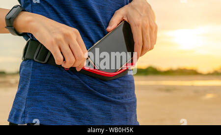 Giovane donna asiatica tenere lo smartphone nel marsupio prima di eseguire cardio esercizio al mattino. Allenamento all'aperto. Runner e banda intelligente dispositivo indossabile. Foto Stock