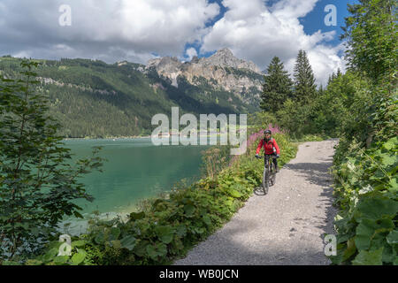 Bella e attiva donna anziana, a cavallo della sua e-mountain bike nella valle di Tannheim lungo il lato Haldensee, Tirolo, Austria, con il villaggio di Tannheim Foto Stock