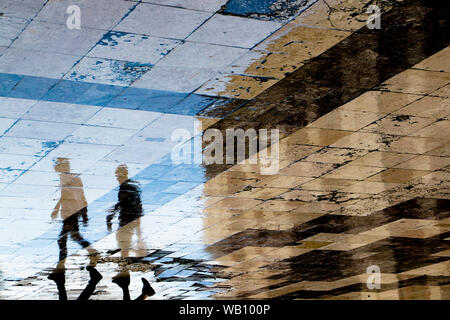 Sfocata riflessione ombra sagome di due giovani uomini a camminare su una pavimentazione di vintage su bagnato via della città, uno che porta una bottiglia e un sac Foto Stock