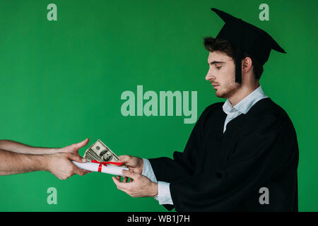 Studente nel cappuccio accademico tenendo diploma con nastro rosso e offrendo corrompere isolato su verde Foto Stock