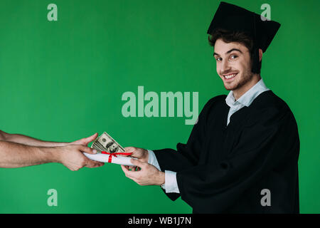 Studente nel cappuccio accademico tenendo diploma con nastro rosso e offrendo corrompere isolato su verde Foto Stock